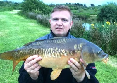 Man holding a large carp fish outdoors near a pond, showcasing a successful fishing catch in a lush green landscape.