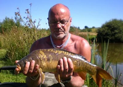 Man holding a large carp fish while standing by a serene lake surrounded by greenery on a sunny day.