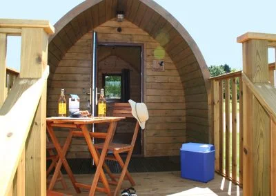 Cozy wooden glamping pod entrance with a small outdoor table, chairs, and cooler, surrounded by nature. Ideal for camping and outdoor adventures.