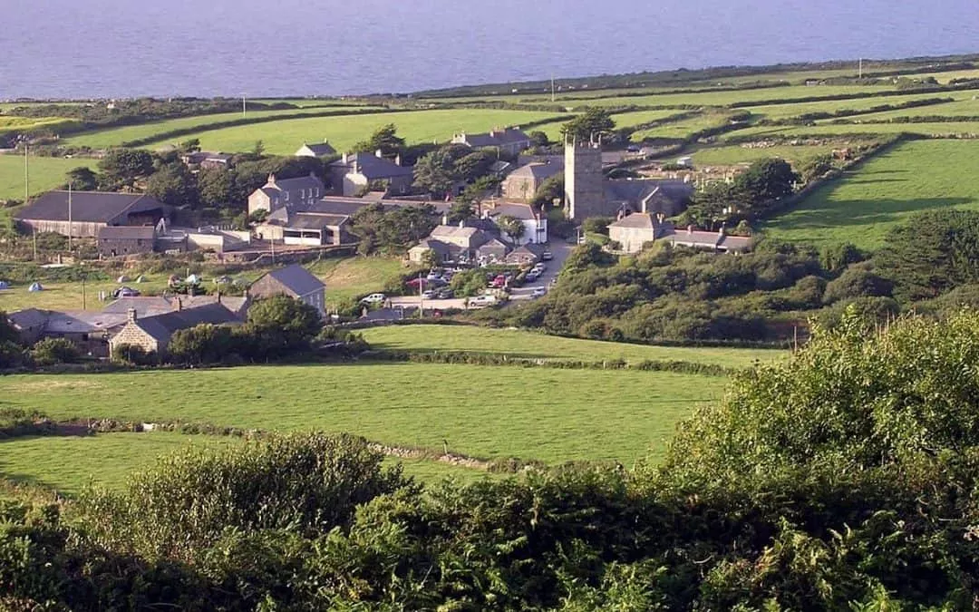 Aerial view of a picturesque village surrounded by lush green fields and rolling hills, showcasing traditional stone houses and a historic tower in the background.